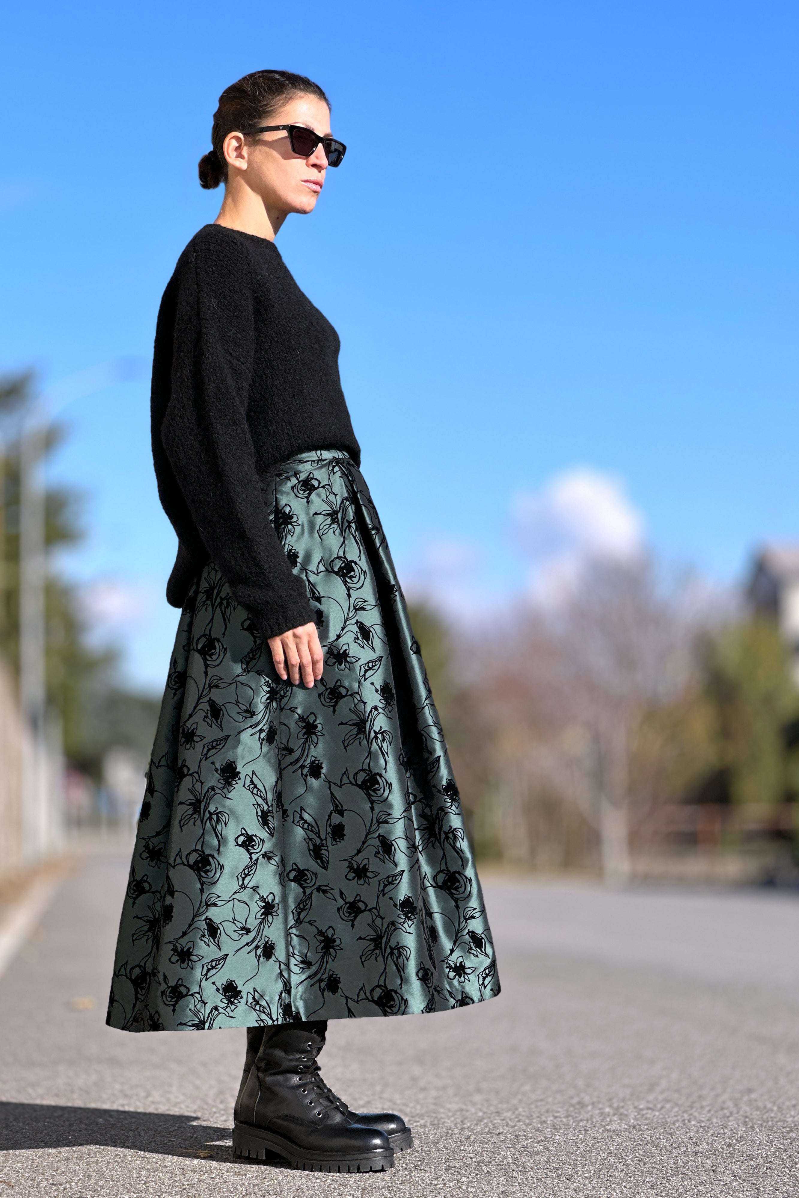 Donna con gonna midi verde Milano a pieghe e disegni floreali, maglione nero e stivaletti neri