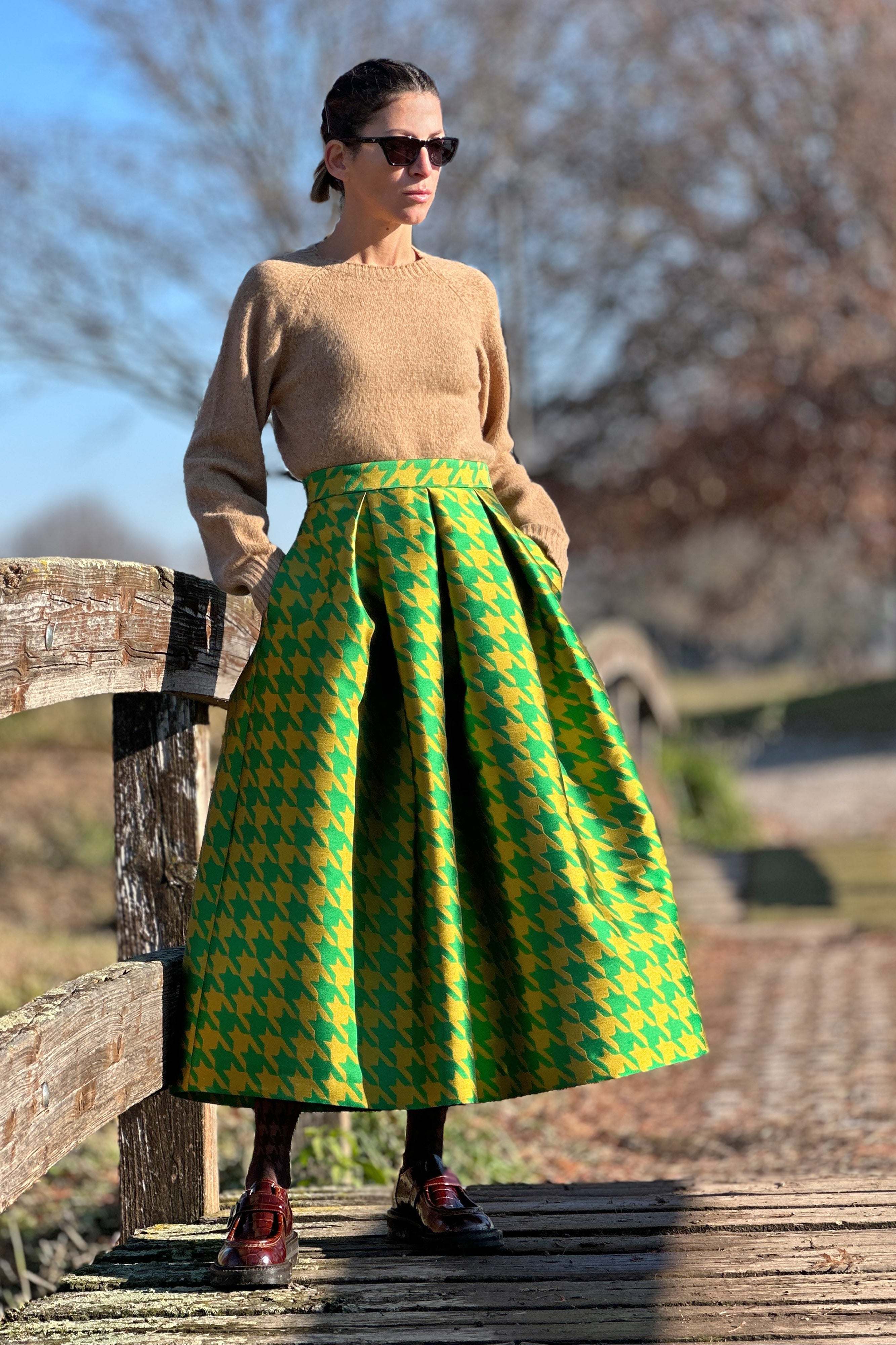 Woman wearing a green and yellow Gonna Pied de poule pleated jacquard skirt standing outdoors on wooden fence background